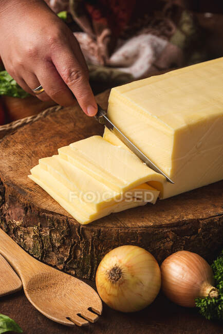 Cropped unrecognizable peron cutting block of cheese with knife on wooden stand near raw onions against organic spatulas — Stock Photo