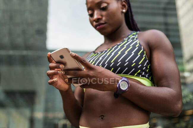 Африканська жінка в спортивній манері переглядає сучасні смартфони, стоячи на розмитому фоні вулиць міста під час тренування на відкритому повітрі. — стокове фото