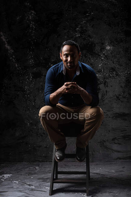 Encantado elegante macho étnico sentado em banquinho de madeira em estúdio no fundo escuro e olhando para a câmera — Fotografia de Stock