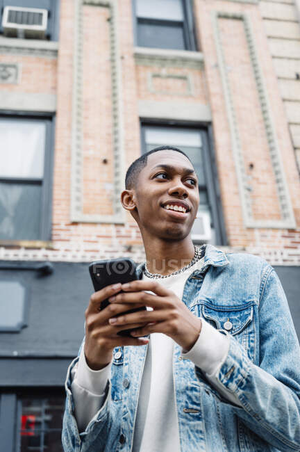 De baixo preto positivo jovem em jeans roupa mensagens no telefone celular enquanto caminhava na cidade olhando para longe — Fotografia de Stock