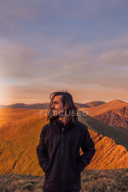 Улыбающийся турист, стоящий на скалистом холме в высокогорье и наслаждающийся видом на горный хребет на закате в Уэльсе — стоковое фото