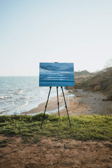 Картина на мольберті розміщена на піщаному пляжі, де миють пінисте море, оточене скелястими скелями в сонячний день — стокове фото