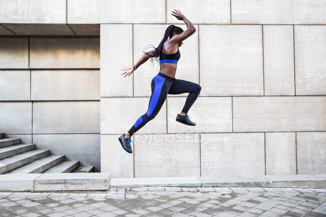 Seitenansicht einer muskulösen Afroamerikanerin in Sportbekleidung, die hoch in der Luft springt, während sie in der Nähe der Mauer eines modernen Gebäudes an der Stadtstraße trainiert — Stockfoto