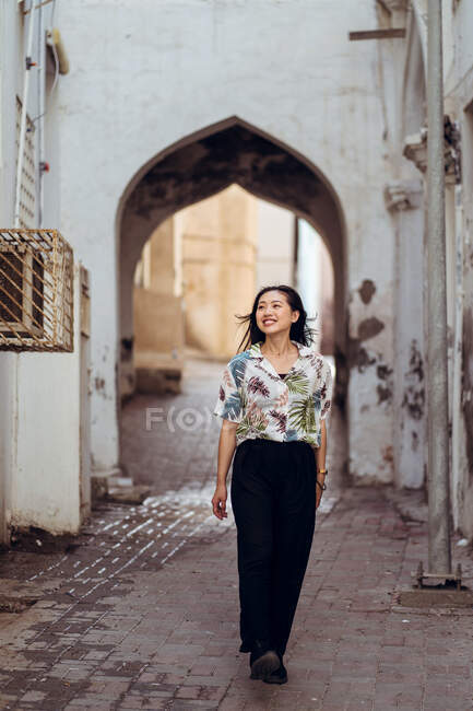Full body delighted young female traveler in casual outfit strolling along narrow street with archway of ancient building while visiting Muttrah Souq marketplace in Muscat province of Oman — Stock Photo