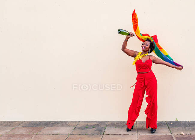 Fröhliche Afroamerikanerin in eleganter Kleidung mit einer Flasche alkoholischen Getränks und einer bunten Fahne, die auf hellem Hintergrund nach oben blickt — Stockfoto