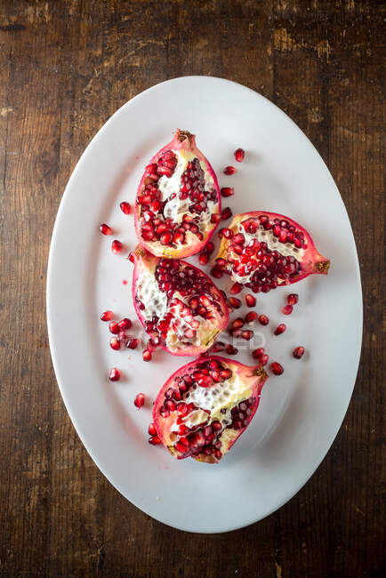 Composición bodegón vista superior con trozos cortados de fruta fresca de granada madura con semillas en plato blanco colocado sobre mesa de madera - foto de stock