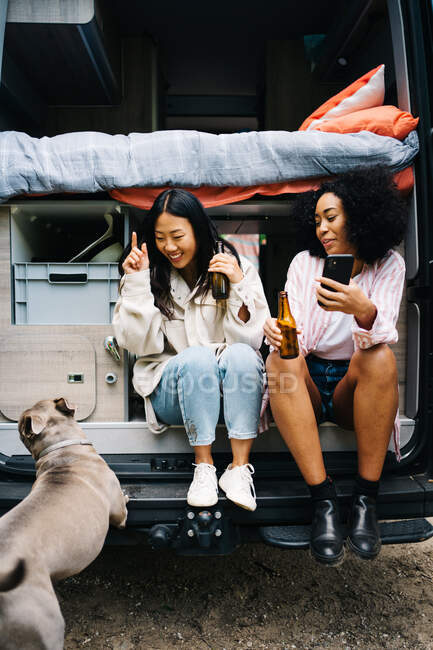 Joyful jovens multiétnicos amigos do sexo feminino beber cerveja e se divertindo enquanto sentado no limiar da van campista perto do cão e desfrutar de férias — Fotografia de Stock