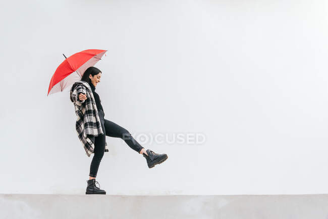 Happy young female with red umbrella walking and balancing on border against gray wall on rainy day on street — Stock Photo