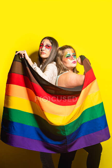 Vue latérale de petites amies multiraciales avec un maquillage créatif démontrant le drapeau LGBT sur fond jaune — Photo de stock