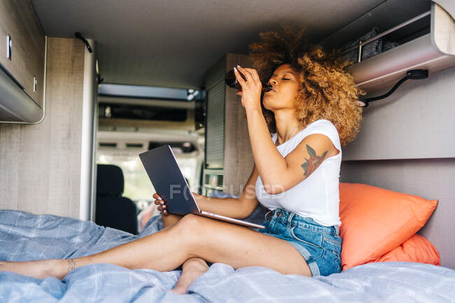 Junge Afroamerikanerin mit lockigem Haar trinkt Bier und schaut während der Sommerferien Filme auf dem Laptop, während sie sich im Wohnmobil ausruht — Stockfoto