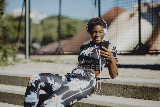 Mujer afroamericana joven en ropa deportiva colorida escuchando música en auriculares mientras está sentada en escaleras de piedra en el parque - foto de stock