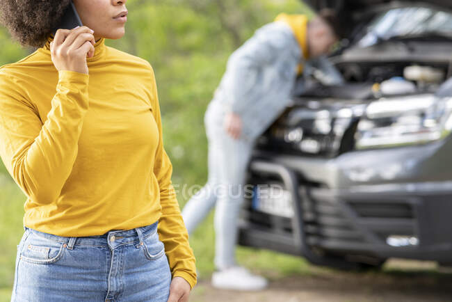 Cropped unrecognizable black woman making call to emergency service while young man trying to fix engine of van during road trip in countryside — Stock Photo