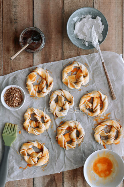 Vista superior de pretzels crudos dulces con mermelada de higo y queso crema colocados en papel cerca de huevo crudo y cepillo sobre mesa de madera - foto de stock