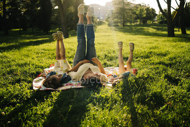 Jovem mulher feliz e irmãs adoráveis em vestidos semelhantes deitado em cobertor na grama verde, enquanto passar o dia de verão juntos no parque e pernas para cima — Fotografia de Stock