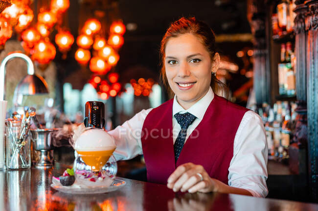 Jovem garçonete experiente usando arma de fumaça de sabor bluster enquanto decora coquetel no balcão do bar — Fotografia de Stock