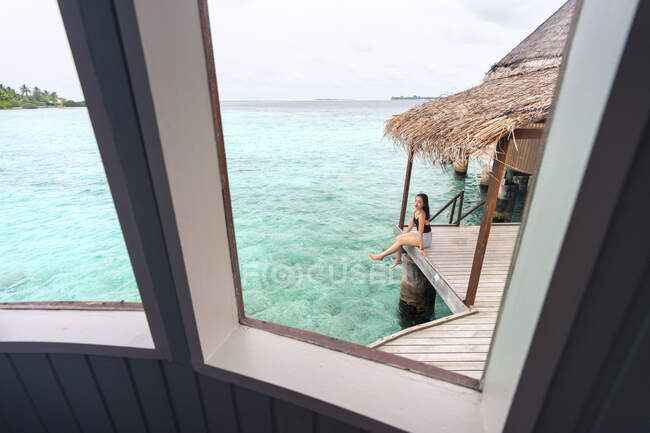 Blick aus dem Fenster auf junge, nachdenkliche Asiatinnen in lässiger Kleidung, die auf einem Holzsteg sitzen und sich auf den Malediven entspannen — Stockfoto