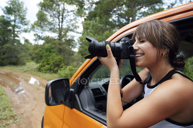 Content female traveler sitting in van and taking pictures of nature in forest on photo camera during road trip in summer — Stock Photo
