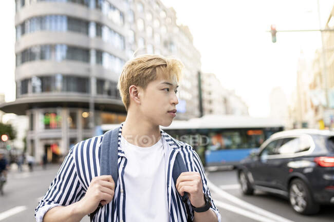 Trendy asiatica modello maschile con capelli biondi e zaino in piedi nella strada della città e guardando altrove — Foto stock