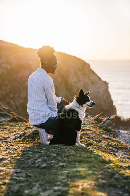 Vista laterale della giovane proprietaria afroamericana con il cane Border Collie che trascorre del tempo insieme sulla spiaggia vicino al mare che ondeggia al tramonto contemplando viste — Foto stock