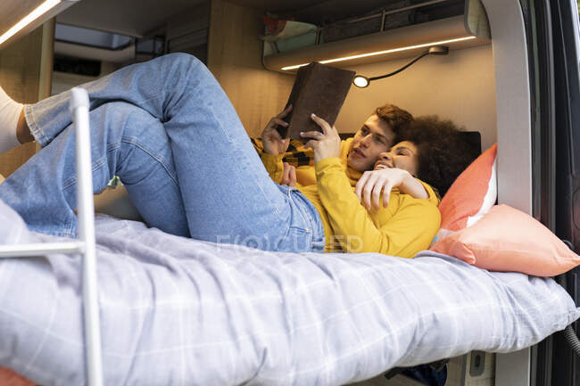 Divers jeunes hommes et femmes embrasser et lire livre intéressant tout en se refroidissant sur le lit en van pendant le voyage sur la route — Photo de stock