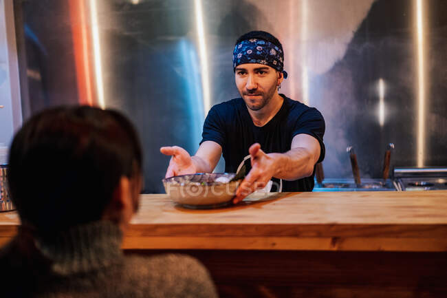 Homme barbu en bandana et t-shirt noir plaçant la plaque avec nouille sur le comptoir en bois pour le client dans ramen bar — Photo de stock