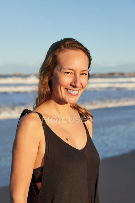 Sonriente hembra en vestido de verano de pie en la orilla del mar de arena y mirando a la cámara - foto de stock