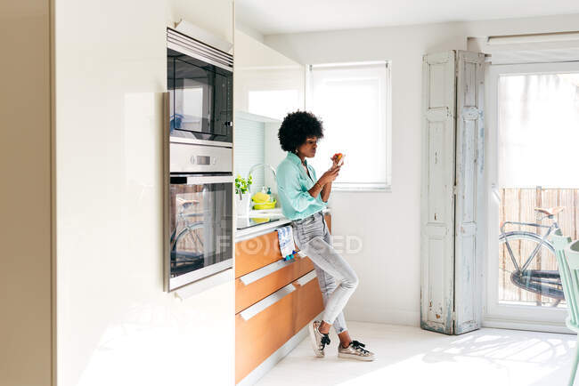 Вид сбоку на молодую афроамериканку в повседневной одежде, которая ест яблоко и просматривает Интернет по мобильному телефону, стоя дома на кухне — стоковое фото