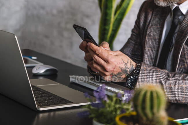 Recorte ejecutivo masculino tatuado irreconocible en mensajería de texto chaqueta a cuadros en el teléfono celular contra el ordenador portátil durante el teletrabajo - foto de stock