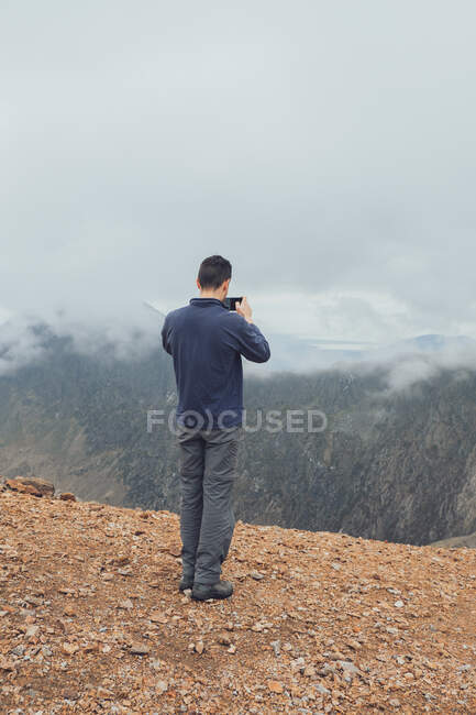 Back view of unrecognizable male hiker standing on hill and taking picture of amazing highlands on smartphone on misty day during vacation in Wales — Stock Photo