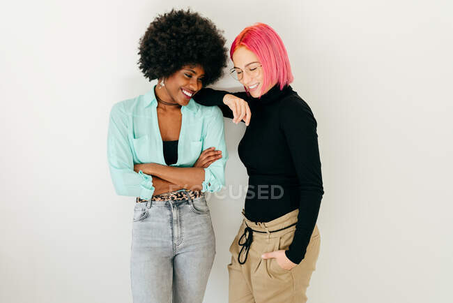 Jovem mulher de cabelos cor-de-rosa alegre e namorada afro-americana em roupa elegante em pé juntos no fundo branco — Fotografia de Stock