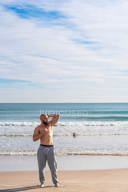 Volle Länge des erwachsenen sportlichen Mannes, der beim Aufwärmen an der sonnigen Küste wegschaut und dabei um seinen Körper herumgeht, mit Hantelscheibe am Band — Stockfoto