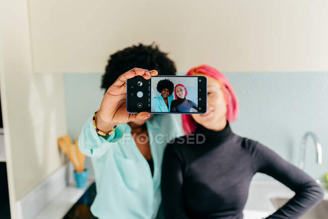 Giovani eleganti e diverse amiche in abiti casual sorridenti mentre si scattano selfie con il telefono cellulare in cucina a casa — Foto stock