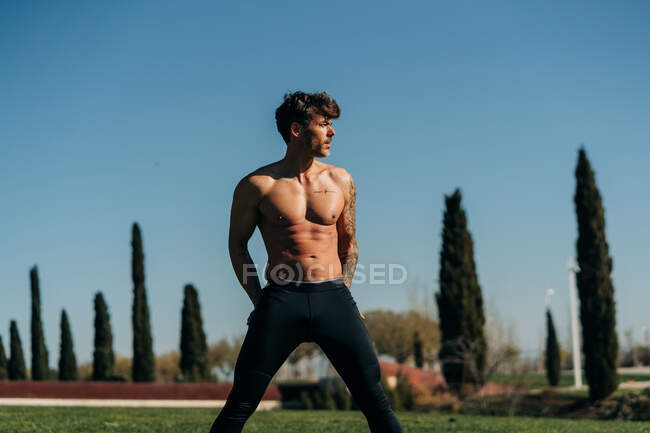 Shirtless male athlete with tattoo and wide legs working out while looking away on meadow — Stock Photo