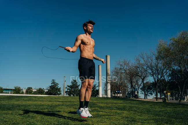 Male athlete in naked torso jumping with skipping rope and looking away on walkway during cardio training in park — Stock Photo