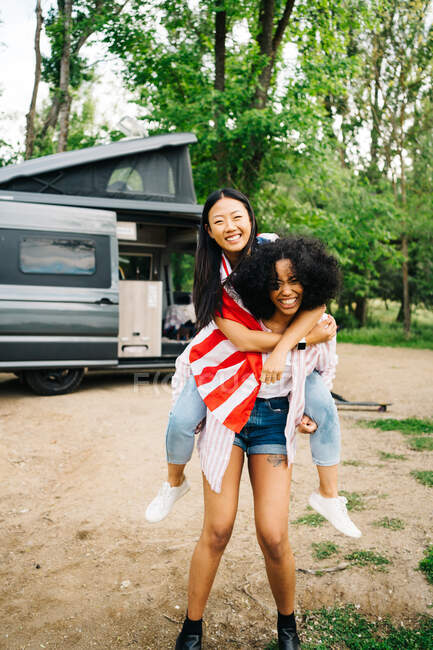 Pleine longueur de jeune femme afro-américaine gaie donnant tour de dos au contenu petite amie asiatique avec drapeau américain tout en profitant de l'aventure estivale ensemble près du camping-car dans la forêt — Photo de stock