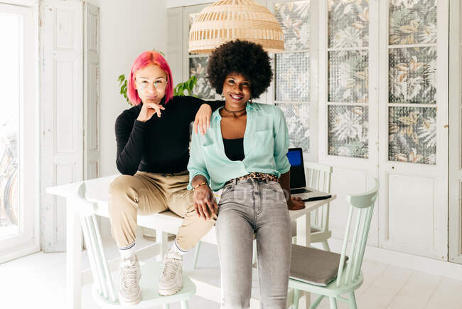 Веселі молоді стильні багаторасові друзі-жінки дивляться на камеру, сидячи разом на столі у світлій кімнаті — стокове фото