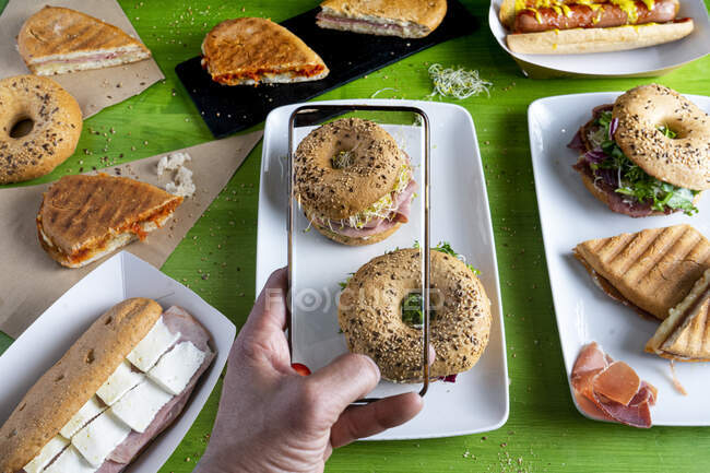De cima de pessoa sem rosto tirando foto de deliciosas sanduíches de bagel usando smartphone com exibição sem moldura — Fotografia de Stock