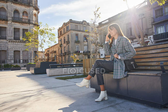 Full length of modern millennial female in stylish spring outfit sitting on bench and answering phone call while resting on urban street — Stock Photo