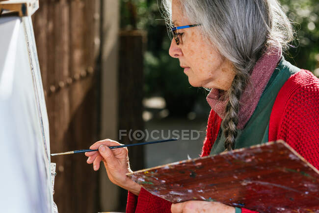Vista lateral de artistas femeninas envejecidas en pintura de máscara protectora sobre lienzo en el patio trasero en el día soleado en verano - foto de stock