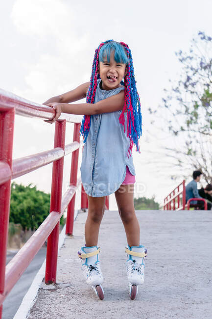 Веселый этнический ребенок с высунутым языком и красочными косичками катается на роликовых коньках, глядя в камеру в городе — стоковое фото