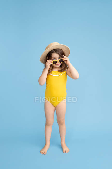 Cuerpo completo de linda niña feliz con traje de baño amarillo y sombrero de paja con gafas de sol con estilo de pie sobre fondo azul y mirando a la cámara - foto de stock
