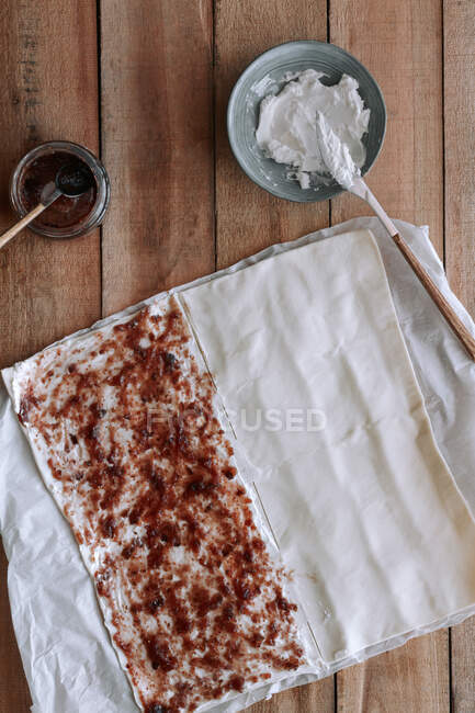 De arriba masa de pastelería delgada cubierta con mermelada de higo dulce y queso crema fresco y colocada en papel sobre una mesa de madera en la cocina - foto de stock