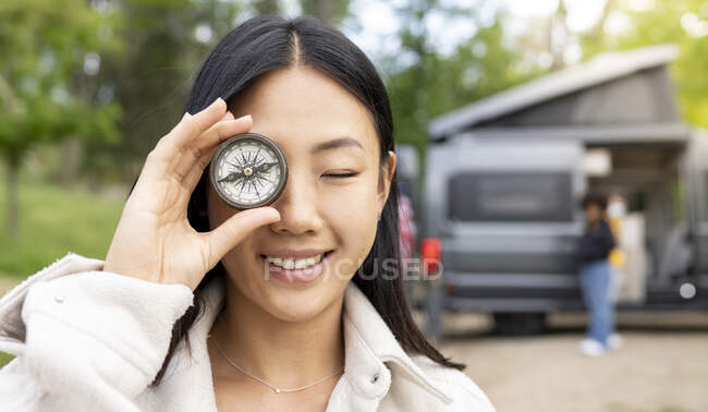 Happy Asian woman smiling and with eyes closed while holding vintage compass near eye during trip in countryside with friends — Stock Photo