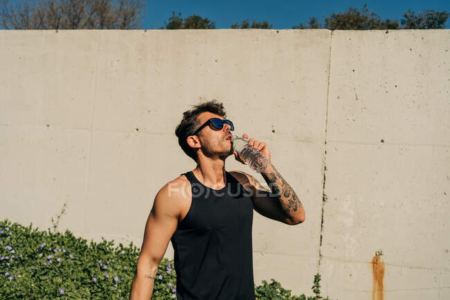 Мужчина-спортсмен с татуировкой в спортивной одежде и солнцезащитных очках пьет воду из бутылки после тренировки на солнце — стоковое фото