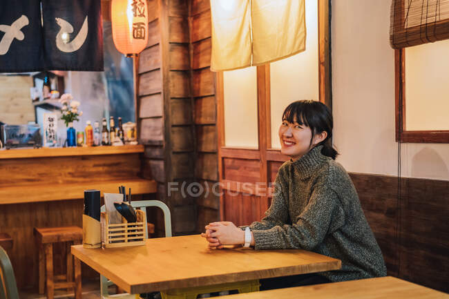 Содержащаяся азиатка в обычном свитере смотрит в камеру с зубастой улыбкой, сидя за деревянным столом в рамен-баре — стоковое фото