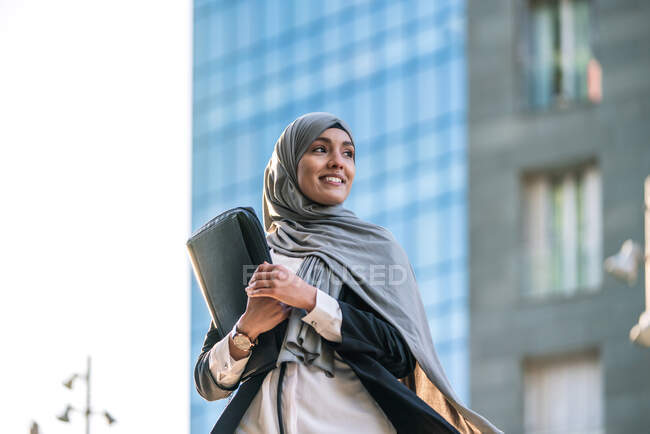 Allegro imprenditore musulmana donna in hijab e con cartella in piedi guardando lontano in strada — Foto stock