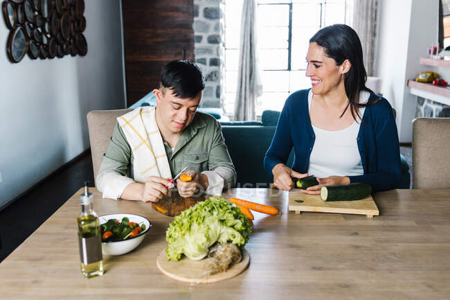 Alegre madre étnica e hijo adolescente con síndrome de Down sentado a la mesa y cortando verduras mientras prepara ensalada para el almuerzo en casa - foto de stock