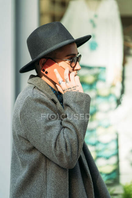 Вид сбоку андрогинного человека в шляпе и современных солнцезащитных очках, говорящего по мобильному телефону, отводя взгляд в сторону стоя на улице при дневном свете — стоковое фото