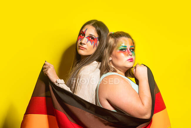 Vista lateral de namoradas multirraciais com maquiagem criativa demonstrando bandeira LGBT contra fundo amarelo — Fotografia de Stock