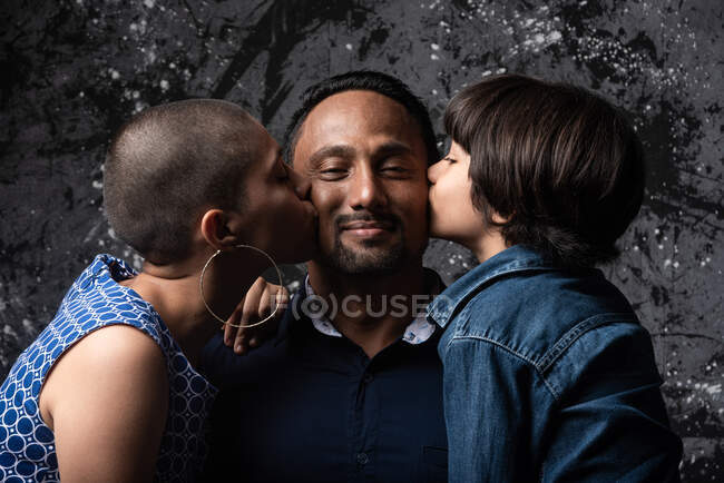 Багатоетнічна любляча жінка і підліток син цілує чоловіка на щоці на темному фоні в студії — стокове фото